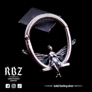 May include: A silver hoop earring with a detailed fairy design. The fairy is sitting on a crescent moon with wings outstretched. The earring is made of solid sterling silver.