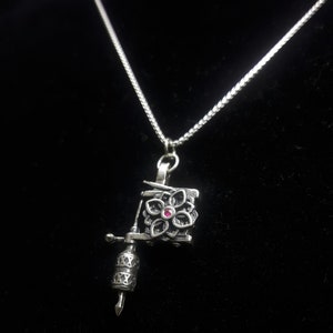 May include: Silver-toned necklace featuring a pendant shaped like a tattoo machine. The pendant has a floral design with a small, red gemstone in the center. The necklace is on a black background.