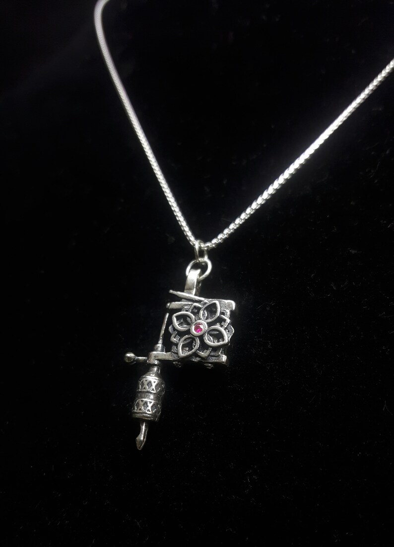 May include: Silver-toned necklace featuring a pendant shaped like a tattoo machine. The pendant has a floral design with a small, red gemstone in the center. The necklace is on a black background.