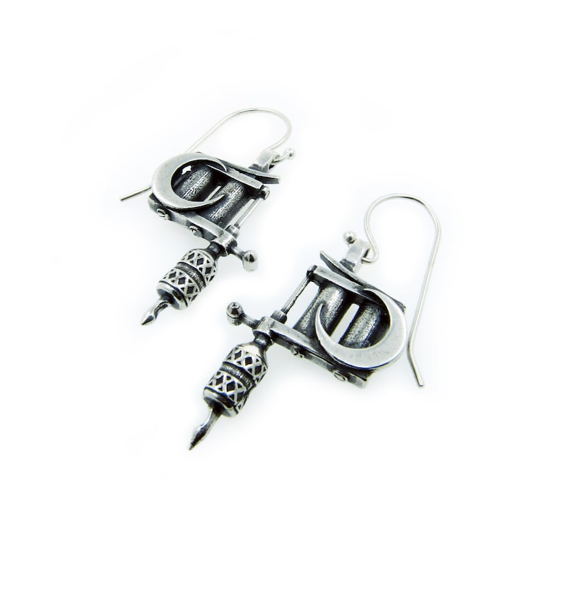 May include: A pair of silver-toned earrings shaped like tattoo machines. Each earring features a curved design, a detailed handle, and a needle-like tip. The earrings hang from a simple hook.