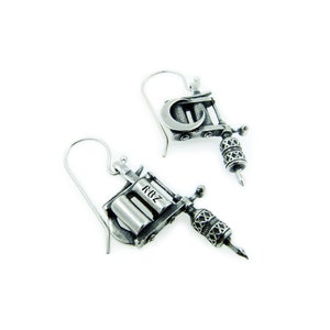May include: Silver-toned metal earrings shaped like tattoo machines. Each earring features a detailed design, including a needle, a barrel, and the letters "ROZ". The earrings hang from a simple hook.