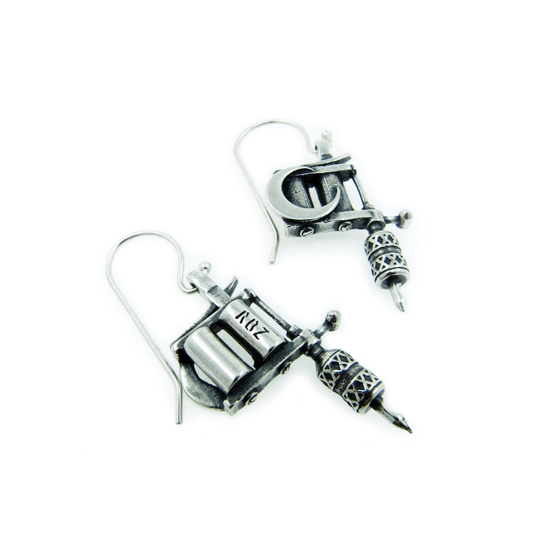 May include: Silver-toned metal earrings shaped like tattoo machines. Each earring features a detailed design, including a needle, a barrel, and the letters "ROZ". The earrings hang from a simple hook.