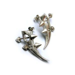 May include: Silver-toned, edgy earrings with a unique design. Each earring features a curved, horn-like shape adorned with multiple cone-shaped spikes. The earrings have a post-and-stud closure, and are set against a white background.