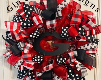 May include: A red, black, and white wreath with a black letter G in the center. The wreath is made of mesh, ribbons, and bows. The ribbons are patterned with polka dots, stripes, and plaid.