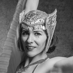 May include: A woman wearing a silver and gold headpiece with a white feather boa. She is wearing a black and white patterned bra and a silver necklace.