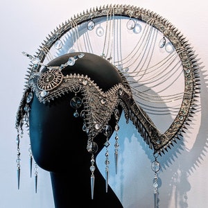 Puede incluir: Un tocado plateado y negro con un marco circular, púas y cadenas colgantes. El tocado está adornado con diamantes de imitación y cuentas.