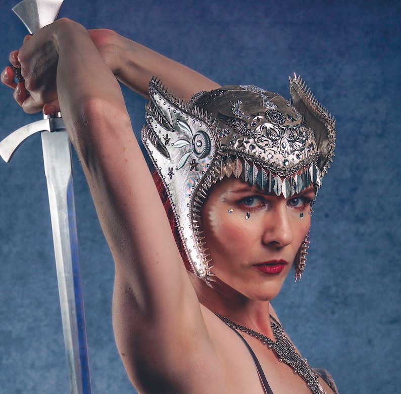 May include: A woman wearing a silver and white helmet with spikes and a sword. The helmet has intricate details and is decorated with rhinestones. The woman is looking at the camera with a serious expression.
