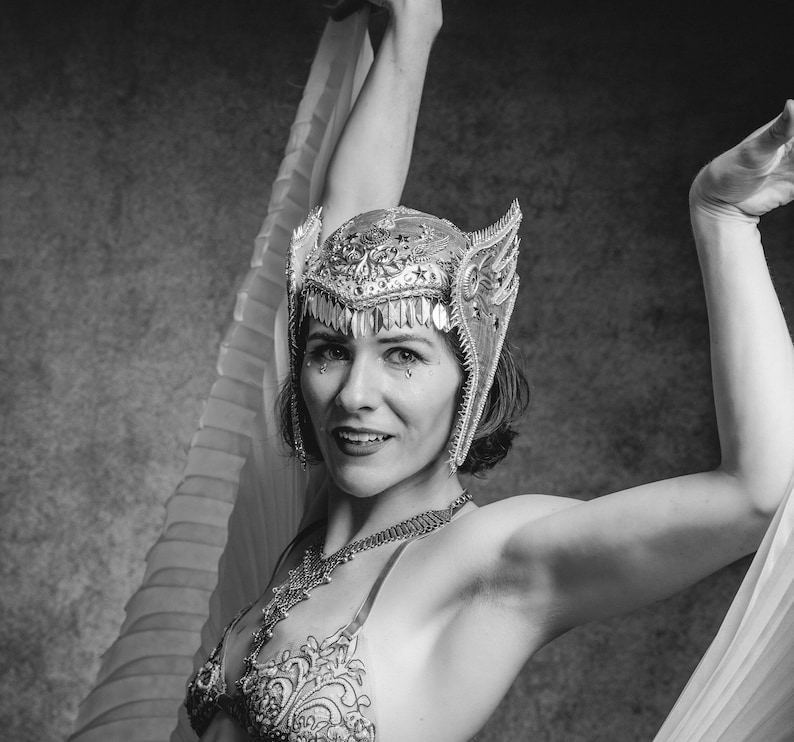 May include: A woman wearing a silver and gold headpiece with a white feather boa. She is wearing a black and white patterned bra and a silver necklace.