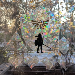 May include: A holographic sticker featuring a silhouette of a video game character holding a sword, with a triangular symbol above. The sticker is on a window with a view of trees and a street.