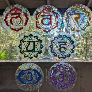 May include: Seven round, colorful chakra window clings with intricate designs and symbols. Each cling features a different color, including red, orange, yellow, green, blue, indigo, and purple. The clings are displayed on a window, with natural light shining through.