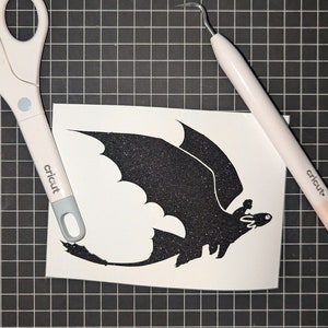 May include: A white vinyl sheet with a black silhouette of a dragon with a rider on its back. The sheet is on a black and white grid surface. A pair of scissors and a weeding tool are also visible.