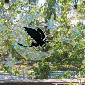 May include: A black silhouette of a dragon flying through a crescent moon, surrounded by iridescent stars. The image is a window sticker with a holographic finish.