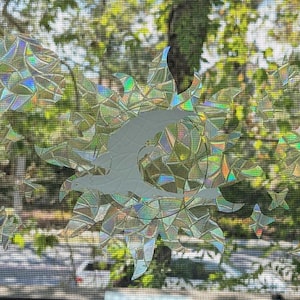 May include: A window with a decorative, iridescent, sun-shaped sticker. The sticker is made of a holographic material that reflects light in a rainbow of colors. The sticker is applied to the window, which is covered with a screen.