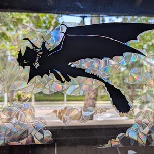 May include: A black dragon silhouette with outstretched wings, flying through iridescent, cloud-shaped decorations. The dragon is a stylized, cartoon-like figure. The background shows a blurred outdoor scene with trees and sunlight.