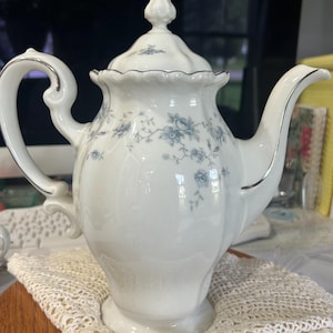 May include: A white porcelain teapot featuring a delicate blue floral pattern, a scalloped rim, and a matching lid with a finial. The teapot has a curved handle and a long spout, both accented with silver trim along the edges of the body and lid.
