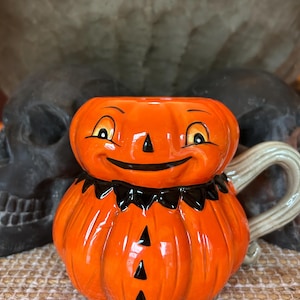 May include: A ceramic mug shaped like a smiling jack-o'-lantern pumpkin. The mug is bright orange with a black collar, black triangular eyes, a black nose, and three black triangular buttons down the front. It features a light beige curved handle.