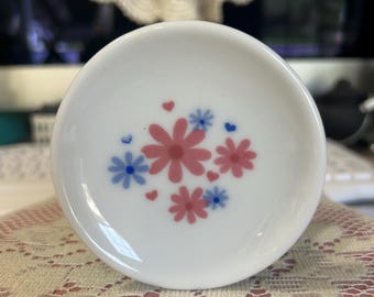 May include: A small, round, white ceramic dish featuring a central floral pattern. The design consists of several pink and light blue daisy-like flowers arranged in a cluster, accented by tiny pink and blue hearts scattered among the petals.