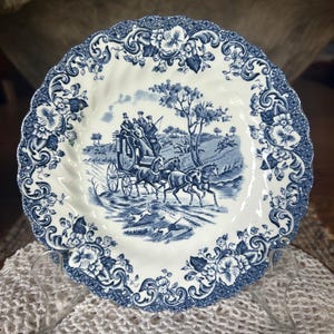 May include: A round ceramic plate with a scalloped edge, featuring a blue and white transferware pattern. The center depicts a horse-drawn carriage with passengers, while the border displays intricate floral scrolls and lattice detailing.