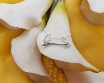 Könnte beinhalten: Eine silberne Pfeilkette mit zwei kleinen silbernen Ringen an der Kette. Die Kette ist auf einer weißen Blume abgebildet.