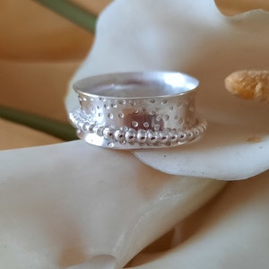 May include: A silver ring with a textured surface and a beaded edge. The ring is sitting on a white flower.