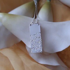 May include: A silver pendant necklace with a rectangular charm featuring a textured design. The charm is hanging from a delicate silver chain.