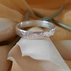 May include: A silver band ring with a textured surface. The ring is on a white and beige background.