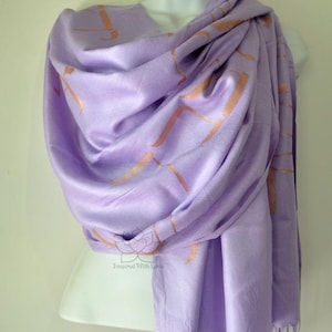 May include: A lavender scarf with a repeating copper-colored geometric pattern is draped over a mannequin. The scarf has a fringed edge and is made of a soft, flowing fabric. The scarf is a light purple color.