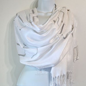 May include: White pashmina scarf with a silver design. The scarf has a fringe border and is made of a soft, lightweight fabric.