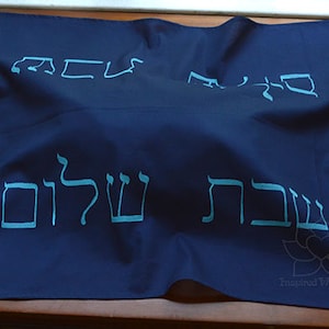May include: A dark blue fabric with Hebrew text in light blue. The text reads "Shema Yisrael" and "Ahava" in Hebrew. The fabric has a white lotus flower design and the text "Inspired With Love" in the bottom right corner.