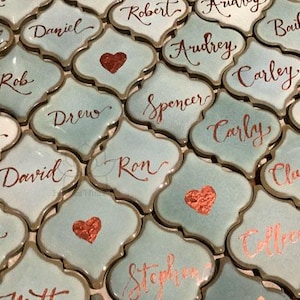 May include: A collection of decorative tiles with names written in calligraphy. The tiles are light blue with gold trim and have a copper heart design.