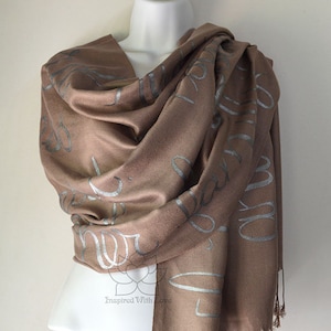 May include: A brown pashmina shawl with a silver print of cursive writing. The shawl is draped over a white mannequin.
