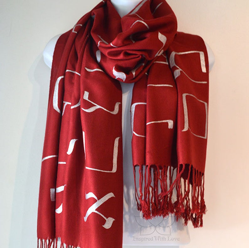 May include: A red scarf with white Hebrew letters printed on it. The scarf has a fringe on the bottom.