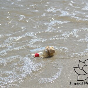 May include: A small glass bottle with a cork top and a red heart inside is partially submerged in the sand on a beach. The bottle is surrounded by foamy water.
