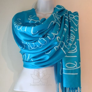 May include: A turquoise blue scarf with white lettering that reads "Inspired With Love". The scarf is made of a soft, lightweight material and has a fringed edge.