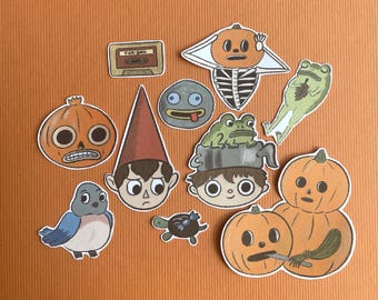 May include: A collection of 11 illustrated stickers featuring characters and objects from the Over the Garden Wall animated series. The stickers include a bluebird, a gnome, a pumpkin, a frog, a cassette tape, and a turtle.