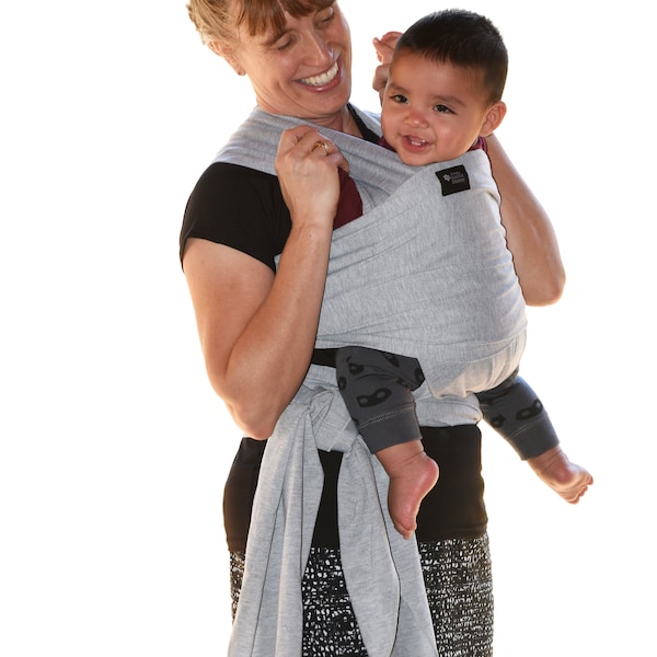 Puede incluir: Una mujer con una falda gris y negra está sosteniendo a un bebé en una envoltura de bebé gris. El bebé lleva una camisa gris y negra con un diseño de máscara negra. La mujer sonríe y mira al bebé.