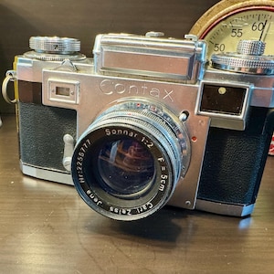 May include: A vintage Contax camera with a silver body and black leather accents. The lens is a Carl Zeiss Sonnar 1:2, f=5cm. The camera is positioned in front of a vintage clock and a yellow can.