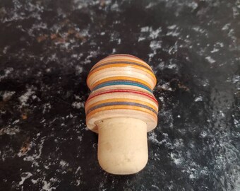 May include: A multi-colored bottle stopper made from recycled skateboard wood. The stopper has a cork base and a layered top with red, yellow, blue, and brown stripes.