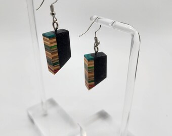 May include: A pair of black and wood earrings with a geometric shape. The earrings are made of layered wood with a black resin coating. The earrings are hanging from silver hooks.