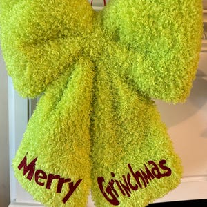 Peut inclure: Un grand nœud vert citron vif fabriqué dans une matière texturée et duveteuse. Les deux pans tombants du nœud portent l'inscription Merry Grinchmas en lettres rouges pailletées, avec Merry sur le pan gauche et Grinchmas sur le pan droit.