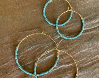 May include: A pair of gold hoop earrings with turquoise blue beads. The earrings are of different sizes, with the larger pair overlapping the smaller pair.
