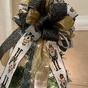May include: A Christmas tree topper bow featuring gold, black, and white ribbons. The bow has a layered design with gold and black patterned ribbons. Some ribbons display a nutcracker soldier design. The bow is placed on top of a Christmas tree.