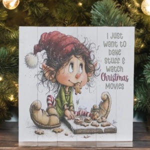 May include: A square wooden sign featuring a cartoon elf in a red hat and green outfit eating a gingerbread cookie. The text reads: I just want to bake stuff & watch Christmas movies. The illustration includes a tray of cookies.