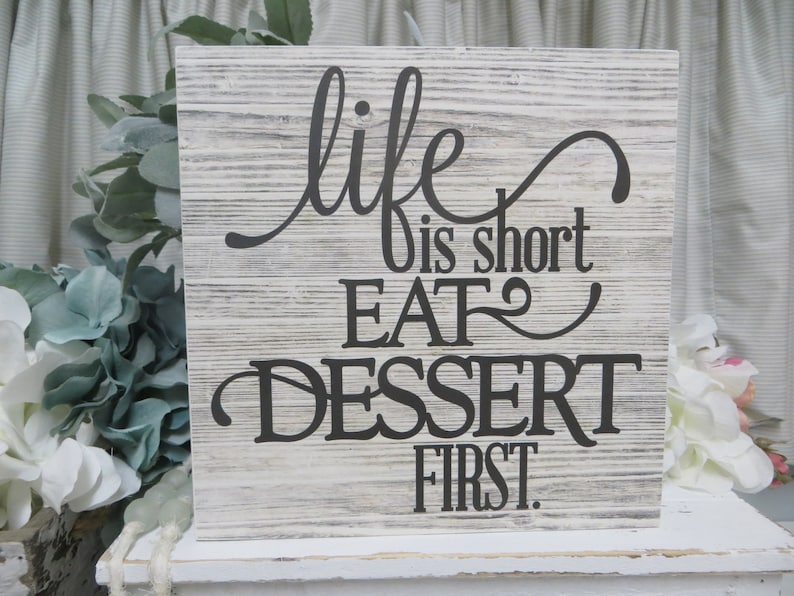 May include: A white wooden sign with black lettering that reads "life is short eat dessert first".