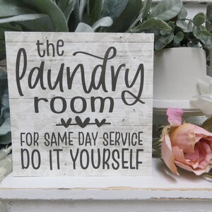 May include: A white wooden sign with black text that reads "the laundry room" with three hearts underneath and "for same day service do it yourself" below. The sign is sitting on a white shelf with flowers in the background.