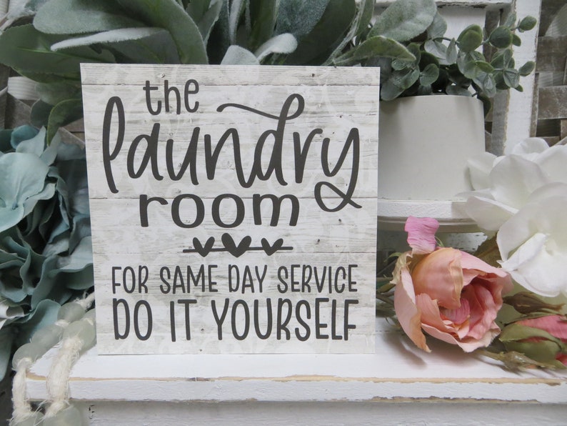May include: A white wooden sign with black text that reads "the laundry room" with three hearts underneath and "for same day service do it yourself" below. The sign is sitting on a white shelf with flowers in the background.