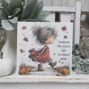 May include: A square decorative sign featuring a whimsical illustration of a child in a red dress walking through fallen leaves and pumpkins. The text reads, Autumn the year's last loveliest smile. The background has a white wood plank pattern.