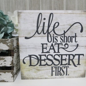 May include: A rustic wooden sign with a whitewashed background and black lettering. The sign reads "life is short EAT dessert first."