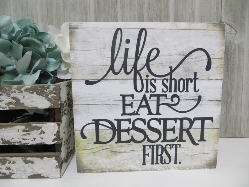 May include: A rustic wooden sign with a whitewashed background and black lettering. The sign reads "life is short EAT dessert first."