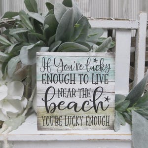May include: A wooden sign with a blue and green background and white painted wood planks. The sign says "If you're lucky enough to live near the beach you're lucky enough".